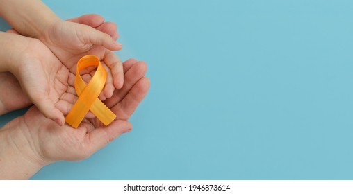 Hands Of Mother And Child Hold Orange Ribbon On Blue Background.Concept. World Multiple Sclerosis Day.Leukemia Cancer Awareness. COPD And ADHD. Layout. Copy Space. Banner. Mock Up.