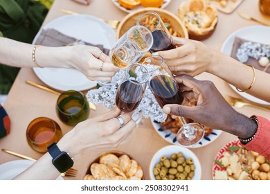 Hands of multi-ethnic group clinking wine glasses in festive gathering celebrating togetherness and diversity while enjoying delicious food and snacks on beautifully arranged table