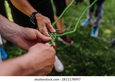 Hands of multiple people work together to tie a green climbing rope knot in an outdoor setting with grass and blurred figures surrounding them