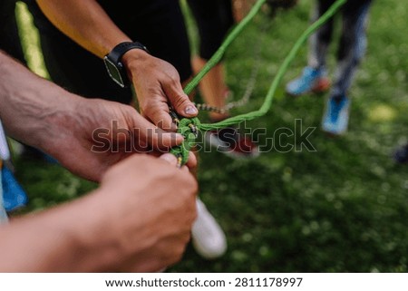 Hands of multiple people work together to tie a green climbing rope knot in an outdoor setting with grass and blurred figures surrounding them