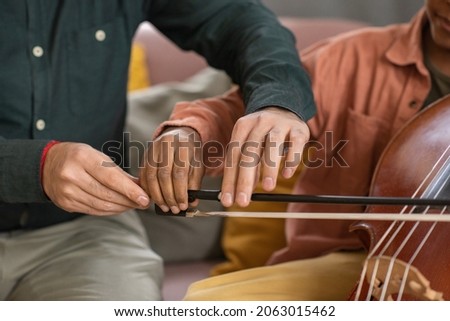 Hands of music teacher and his pupil playing cello while young man holding fiddlestick and helping little boy to glide it