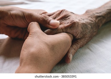 Hands of the old man and a young man on a white bed in a hospital.
