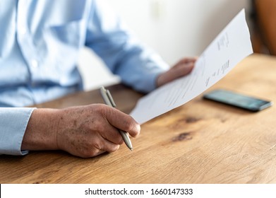 Hands Of An Older Man With Light Blue Shirt, Sitting At A Wooden Table With A Smartphone On It, While He Is Reading A Letter With A Pen In His Hand.