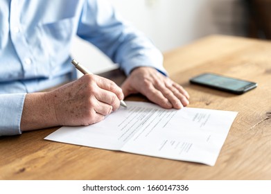 Hands Of An Older Man With Light Blue Shirt, Sitting At A Wooden Table With A Smartphone On It, While He Is Signing A Letter.