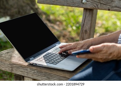 Hands Of An Older Woman Using A Laptop With A Black Screen On The Bench, In A Park. Space For Text.