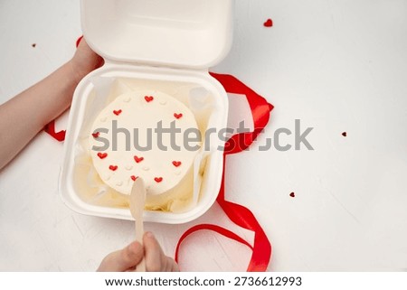 Hands opening a small bento cake decorated with red hearts and Volim te (I love you) written in icing, presented in a takeaway box with a wooden spoon and red ribbon, celebrating love
