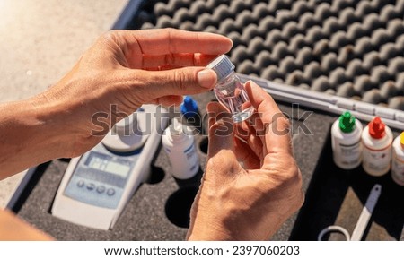 Hands opening a small empty testing bottle with color-coded reagent bottles and digital device in a case in background