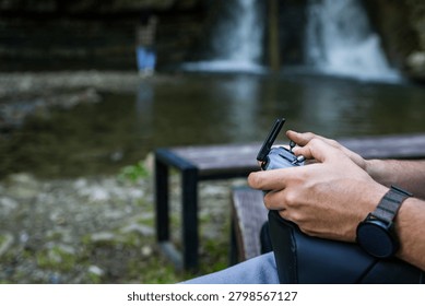 Hands operate a drone controller with a blurred figure near a distant waterfall. The riverbank and pool set a calm backdrop as the pilot manages flight during a relaxed outing