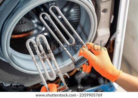 Hands in orange gloves hold two heating elements for a washing machine: one after a long time of use and the other new, against the background of a washing machine.