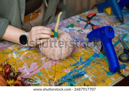 hands with painted nails decorating the stem of a pale ridged pumpkin with green accents at a rustic workshop table with a blue glue gun, concept of diy craft, fall workshop, creative hobby