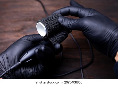 the hands of a permanent makeup artist in black sterile gloves holds a tape in his hands to protect the tattoo machine and wraps it around