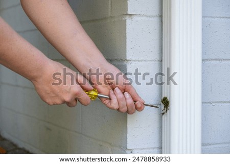 Woman’s hands with Phillips head screwdriver to remove a rusted screw holding a rectangular white metal rainwater downspout to a bracket attached to a painted brick wall
