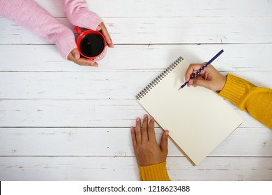 Hands in pink sweater holding red coffee cup with hand in mustard yellow sweater holding polka dot pencil to write some text or messages on blank white writing paper space e.g. New Year’s Resolutions