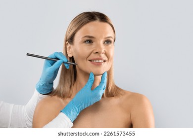 Hands of plastic surgeon wearing blue medical gloves drawing contours with black pen around female patient face before surgery, middle aged blonde woman getting beauty treatment, copy space
