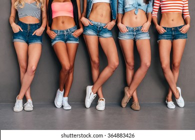 Hands in pockets. Close up of five women wearing jeans shorts and holding hands in their pockets while standing against grey background