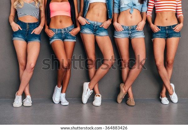 Hands in pockets. Close up of five women\
wearing jeans shorts and holding hands in their pockets while\
standing against grey\
background