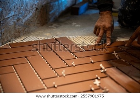 Hands position brown rectangular tiles in a herringbone pattern on a floor. White spacers ensure even spacing. Unfinished OSB walls are visible in the background.
