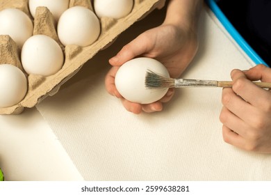 Hands preparing to paint an Easter egg with a brush, surrounded by a carton of white eggs on a clean white surface. Perfect for festive crafts and creative holiday activities.