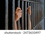 hands of a prisoner behind prison bars on black background
