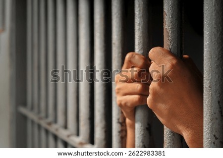 hands of a prisoner behind prison bars on black background	