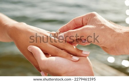 Hands, proposal and engagement ring at lake outdoors for couple, marriage and wedding. Commitment, love and jewelry of woman and man proposing and putting band on finger for trust, care and romance.