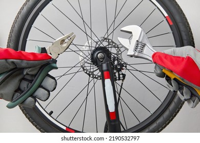 Hands In Protective Gloves With Adjustable Wrenches And Pliers On The Background Of A Bicycle Wheel. Bicycle Repair At Home.