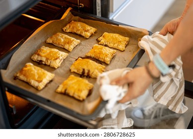 Hands remove baking sheet filled with golden brown pastries from a hot oven.
