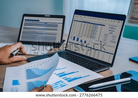 Hands reviewing financial chart document with laptop displaying spreadsheet dashboard and tablet screen, representing data analysis workflow, reporting process, business performance tracking, and digital financial management.