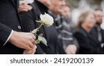 Hands, rose and a person at a funeral in a cemetery in grief while mourning loss at a memorial service. Death, flower and an adult in a suit at a graveyard in a crowd for an outdoor burial closeup
