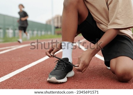 Hands of schoolboy tying black shoelace on gumshoe while standing on squats on race track against his classmates running along stadium