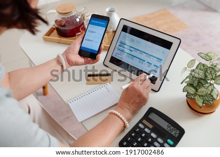 Hands of senior woman checking utility bill on screen tablet computer and paying with banking application on smartphone