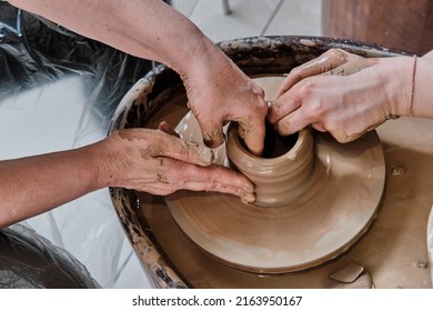 Hands Of Senior Woman And Hands Of Young Girl Sculpting Clay Vase On Potter's Wheel At Pottery Training Lesson. Craftswoman Teaching A Retired Pottery Craft. Hobby Concept