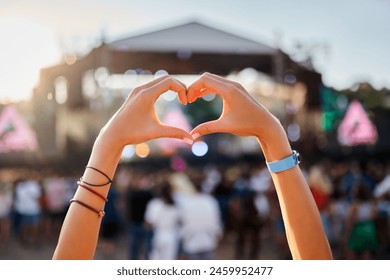 Hands shape heart sign at sunset beach music fest, crowd enjoys live concert. Outdoor summer event, happy fans party, love symbol, festive vibe by the sea. Silhouette, sun glow, entertainment.