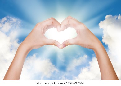 Hands Show A Heart Sign Against A Blue Sky With Clouds With The Sun In The Center. The Concept Of Love And Friendship, Harmony, Protection Of Nature And The Environment. I Love You.