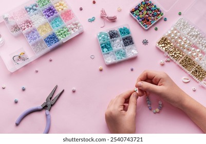 Hands skillfully string beads to make jewelry while surrounded by a variety of beads and crafting tools, set against a soft pink background.