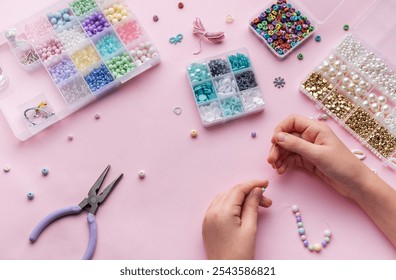 Hands skillfully string beads to make jewelry while surrounded by a variety of beads and crafting tools, set against a soft pink background.