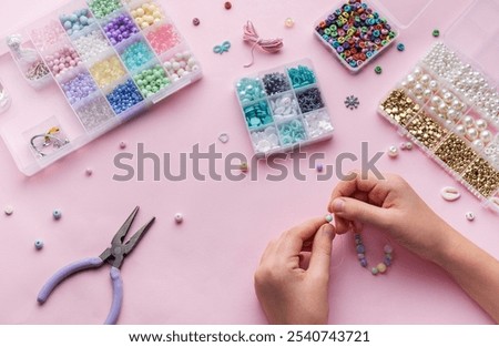 Hands skillfully string beads to make jewelry while surrounded by a variety of beads and crafting tools, set against a soft pink background.
