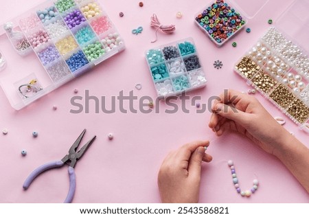 Hands skillfully string beads to make jewelry while surrounded by a variety of beads and crafting tools, set against a soft pink background.