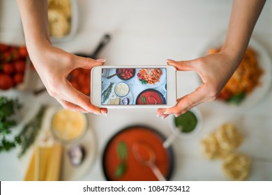 Hands With The Smart Phone Pictures Of Meal. Young Woman, Cooking Blogger Is Cooking At The Home Kitchen In Sunny Day And Is Making Photo At Smartphone. Instagram Food Blogger Workshop Concept.