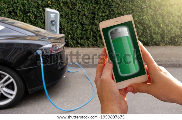 Hands with smartphone on a background of a car at\
the charging station for electric vehicles. Battery level on the\
phone screen