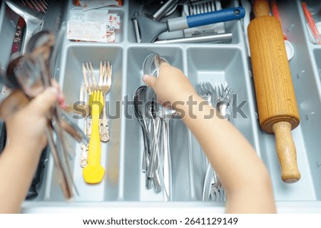Hands are sorting through a kitchen drawer, separating various utensils like forks, spoons, and knives. The drawer is filled with a mix of metal and plastic cutlery.