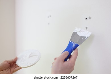 Hands with a spatula and putty against the background of a white wall, a close-up. Scatter holes on the wall - Shutterstock ID 2256637267