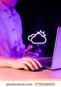 Hands at table with computer and phone featuring illuminated cloud icon in neon purple setting.