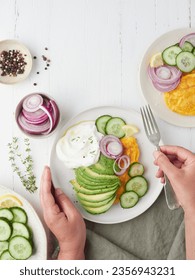 hands to the table holding a fork over plate with avocado, cucumber salad, red onion and omelet. Yogurt sauce seasoned with thyme, lemon and black pepper.