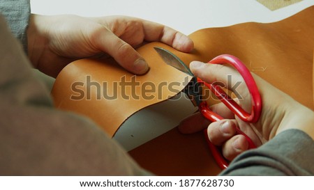 The hands of a tailor make a precise and even cut of a piece of leather with scissors for making a hand-made product for sale.