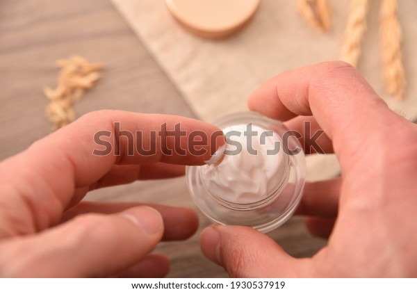 Hands taking natural moisturizing cream of oat\
extract for application with wooden table background with oat\
spikes top view