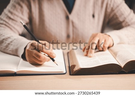 Hands taking notes in a notebook while studying an open Bible, finger tracking the text, warm indoor light and soft focus, conveying faith, devotion, learning and reflection at home.