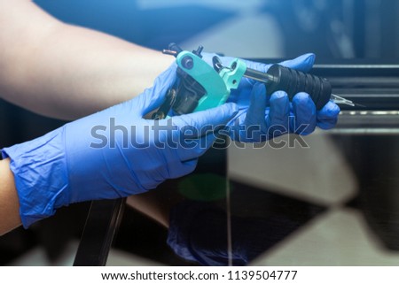 Hands of a tattoo artist in blue rubber gloves hold a blue tattoo machine. Tattoo salon theme