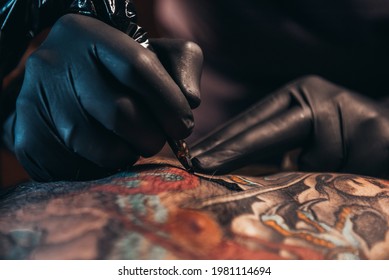 Hands of a tattoo artist wearing black gloves and holding a machine while creating a picture on a man back and the ink is dripping - Shutterstock ID 1981114694