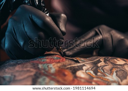 Hands of a tattoo artist wearing black gloves and holding a machine while creating a picture on a man back and the ink is dripping